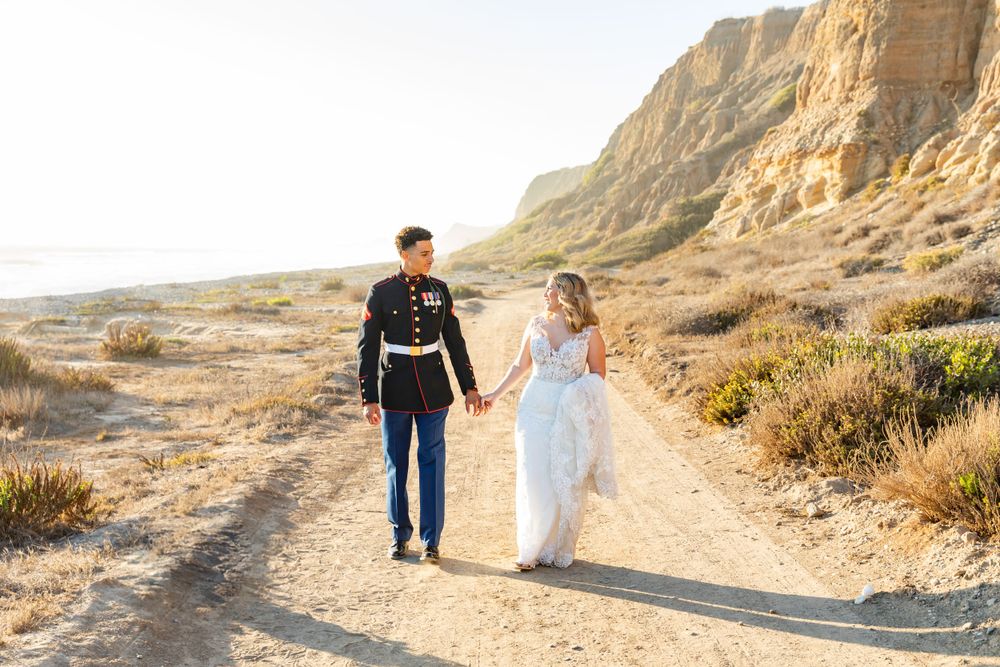 Newlywed couple in military dress blues and white wedding gown walking along desert path during sunset.