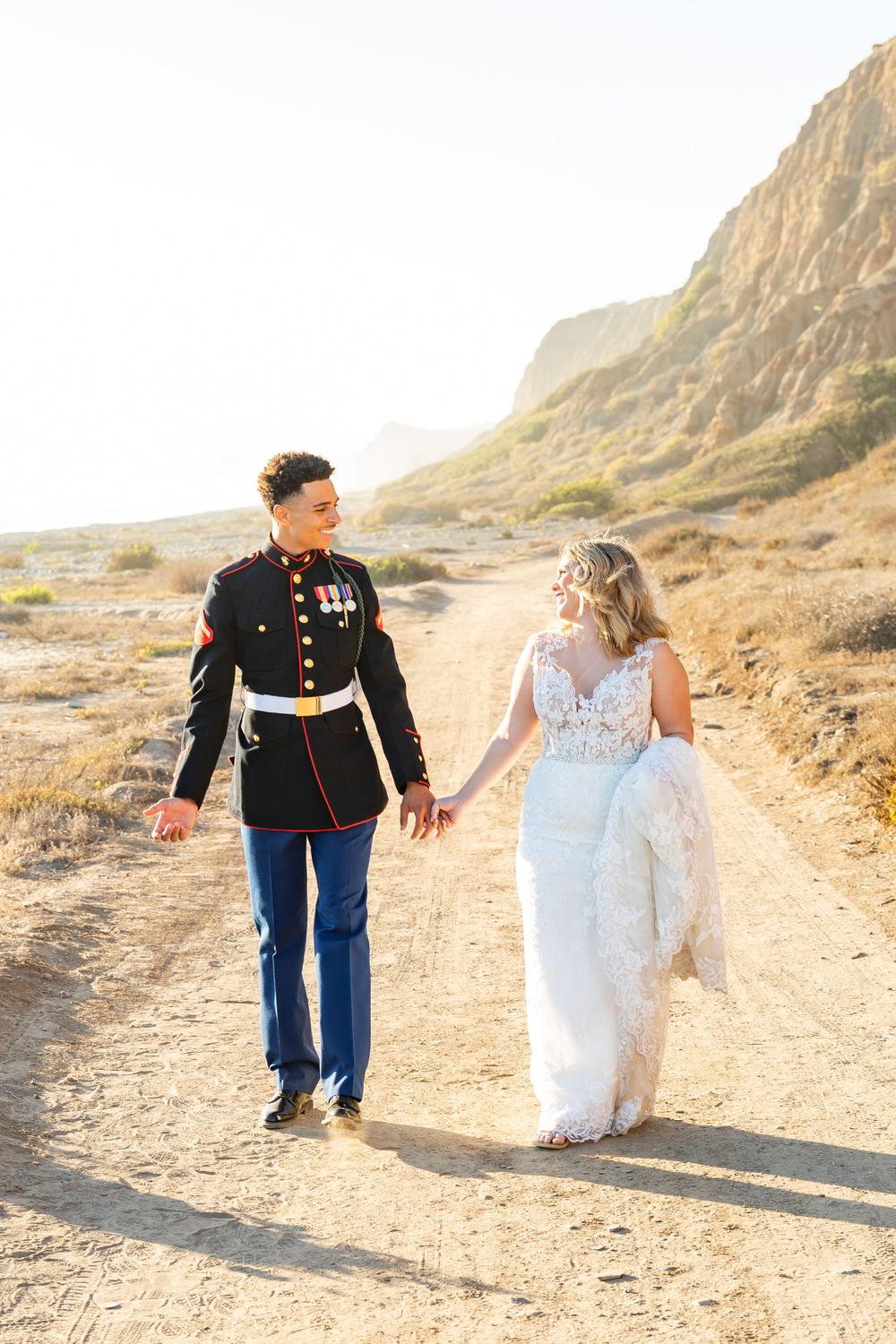 Military wedding couple strolls along scenic desert trail with dramatic cliffs in golden light.
