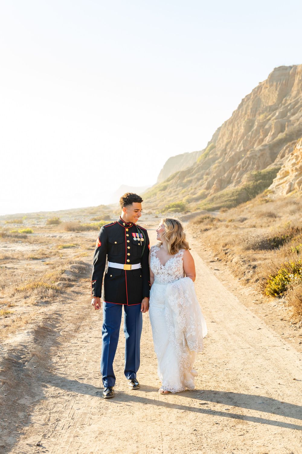 Wedding couple in military uniform and lace dress walking together on desert trail at sunset.