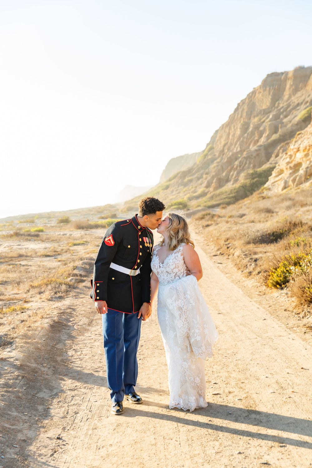 Military couple in dress blues and wedding gown share intimate moment on desert path.