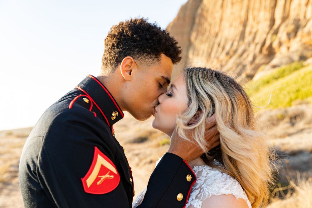 Sunlit wedding portrait of a military couple sharing a romantic kiss against desert cliffs.