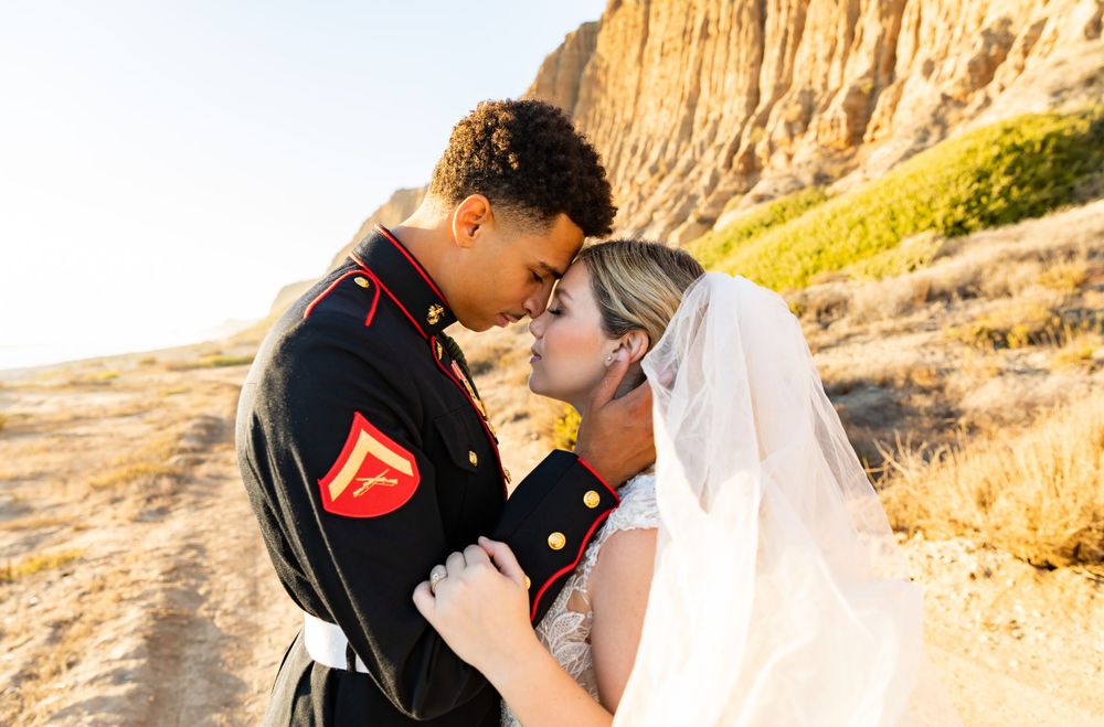 A bride in white and Marine in formal dress uniform embrace during sunset by towering cliffs.