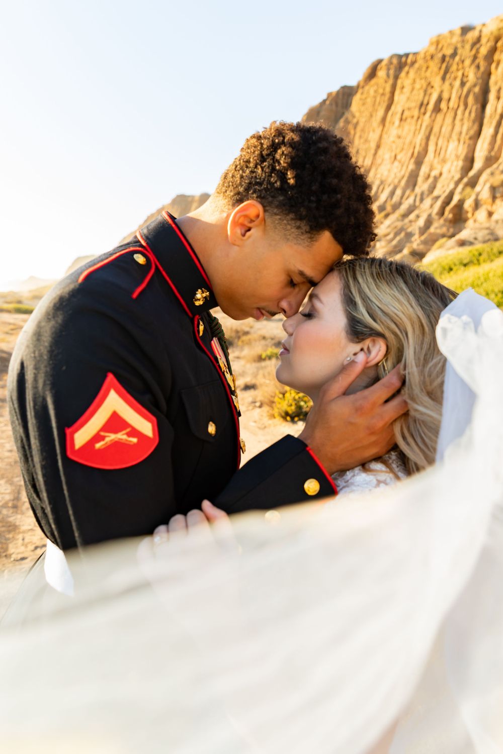Intimate wedding portrait with flowing veil as couple embraces near golden cliffs.