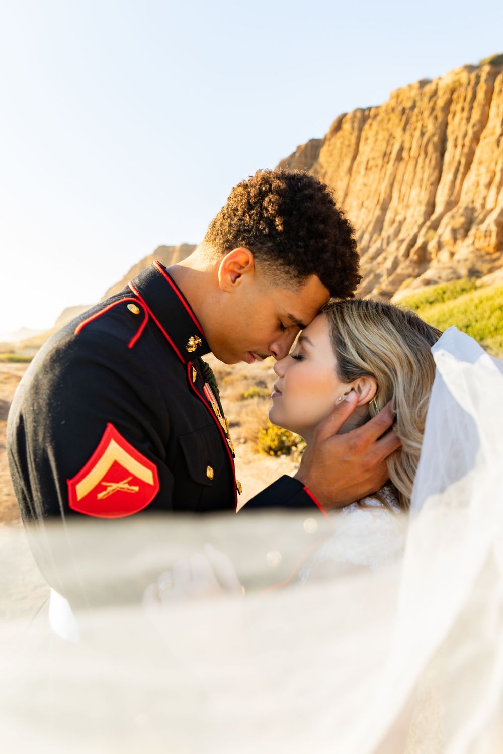 Sun-drenched wedding portrait captures tender moment between military couple with flowing veil.