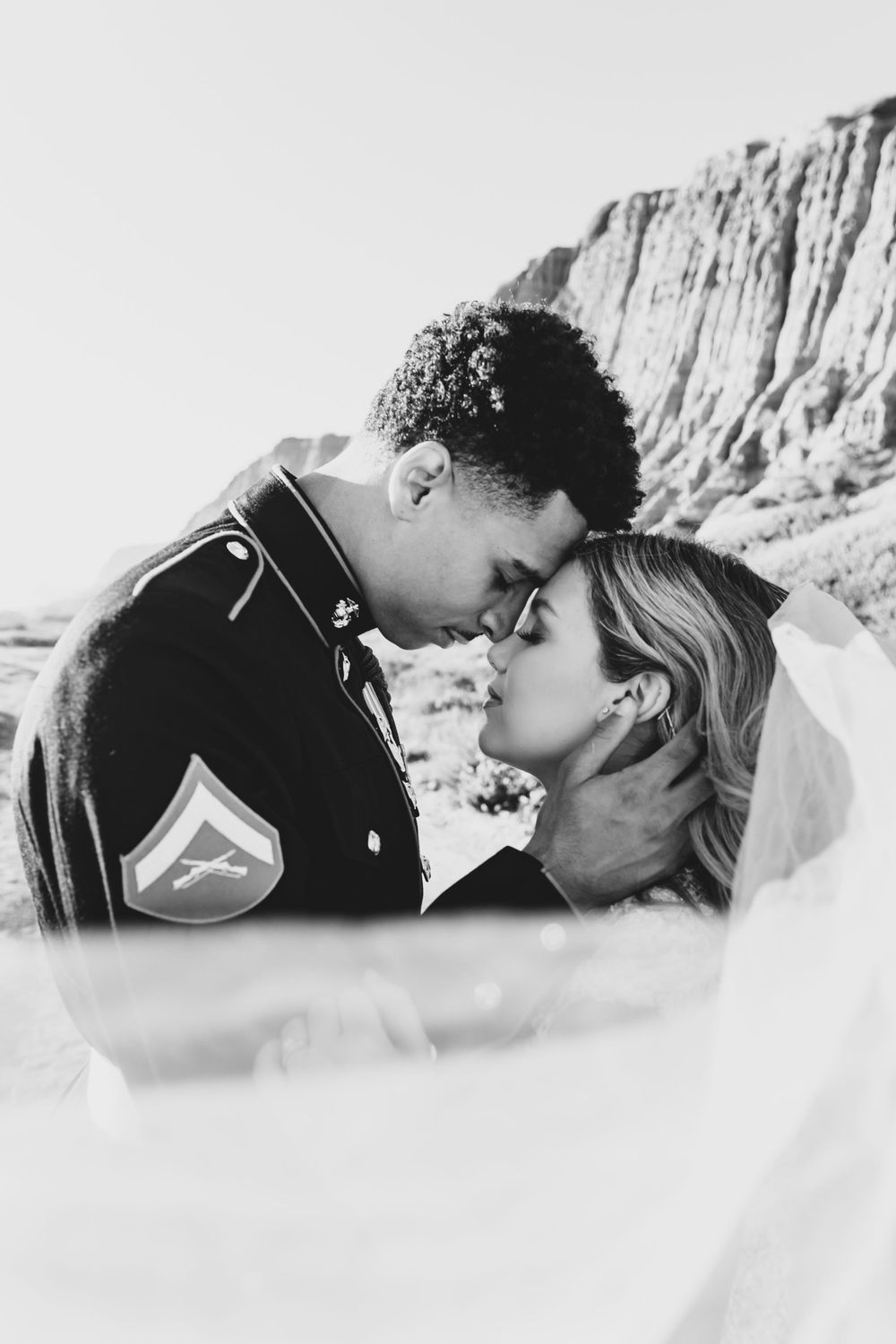 Black and white wedding portrait showing intimate moment between military couple with veil detail.