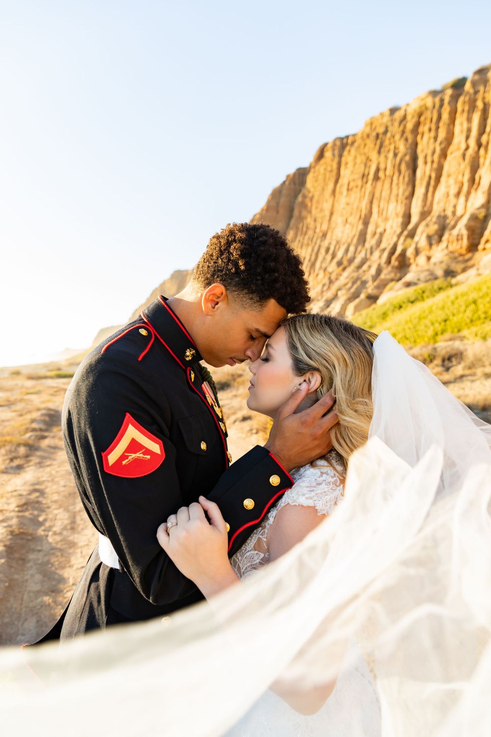 Sunlit wedding photo of military couple sharing romantic moment with flowing veil against cliffs.