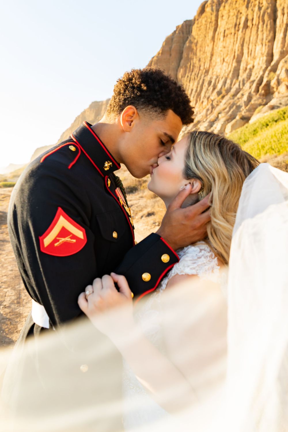 Wedding portrait showing military groom and bride in tender embrace with sunlit cliffs behind.