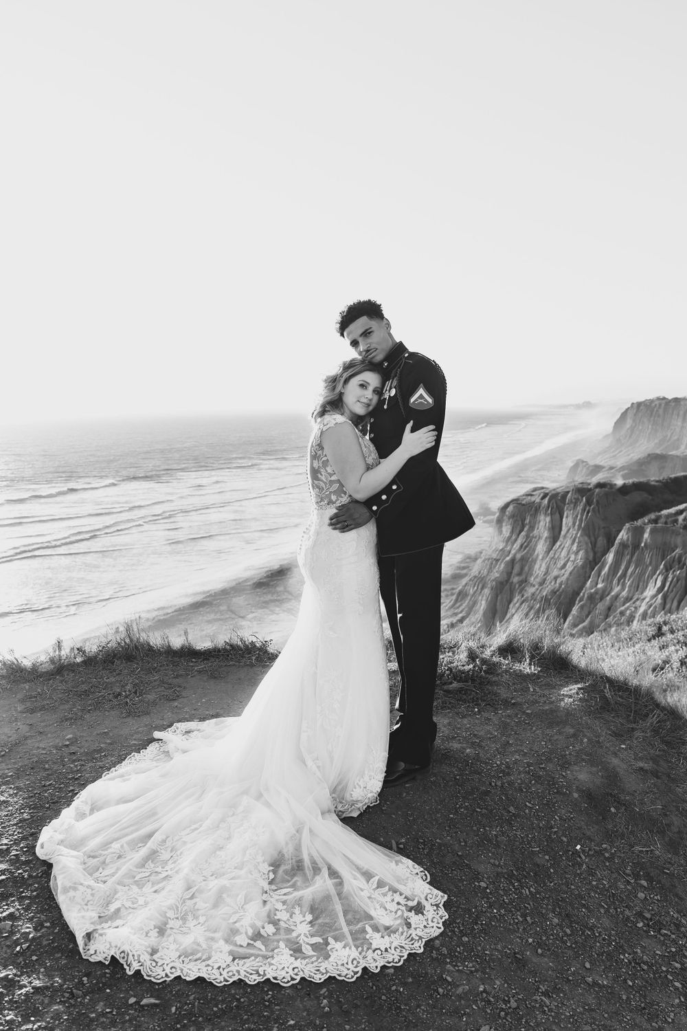 A wedding couple embraces on coastal cliffs overlooking the ocean in dramatic black and white.