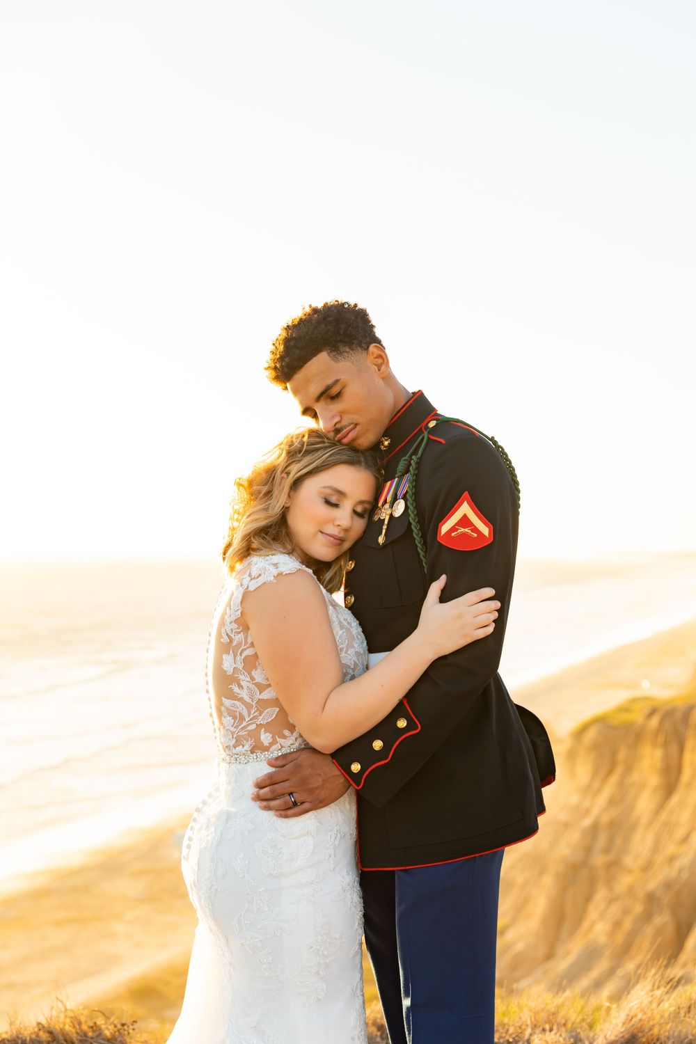 Military wedding couple shares tender moment on clifftop overlooking ocean at sunset.