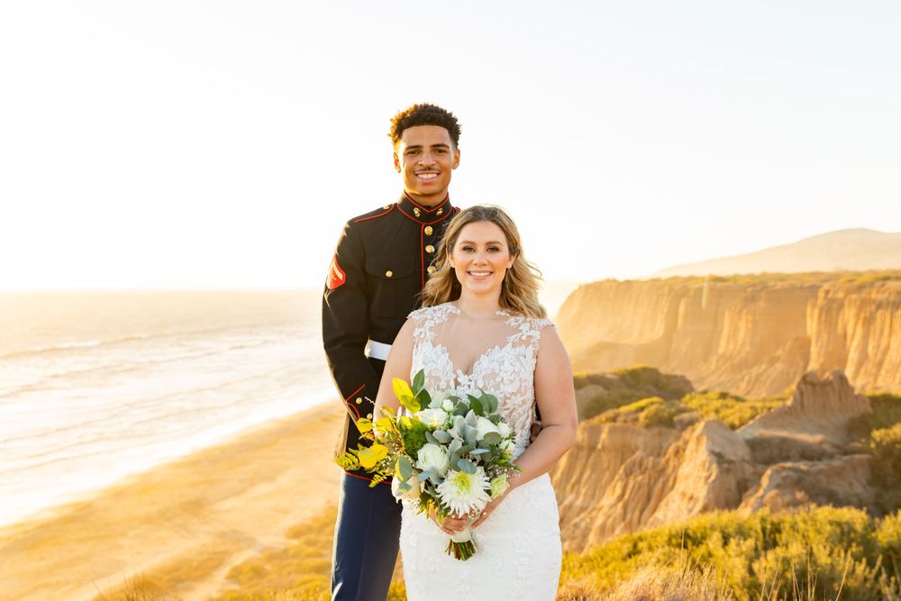 Bride holds yellow and white bouquet while posing with military groom on ocean cliffs.