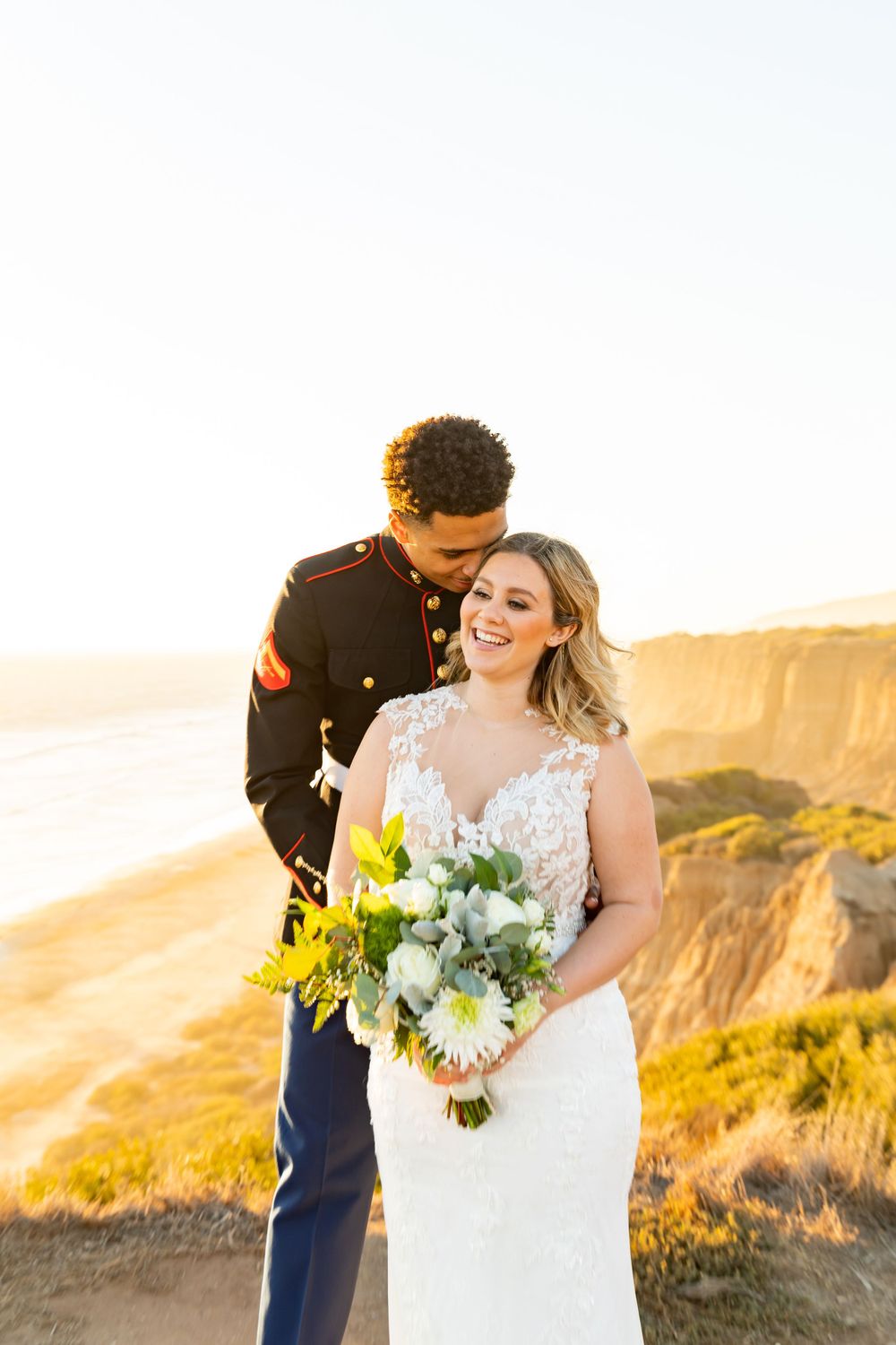 Happy wedding couple shares joyful moment on coastal cliffs with bridal bouquet.
