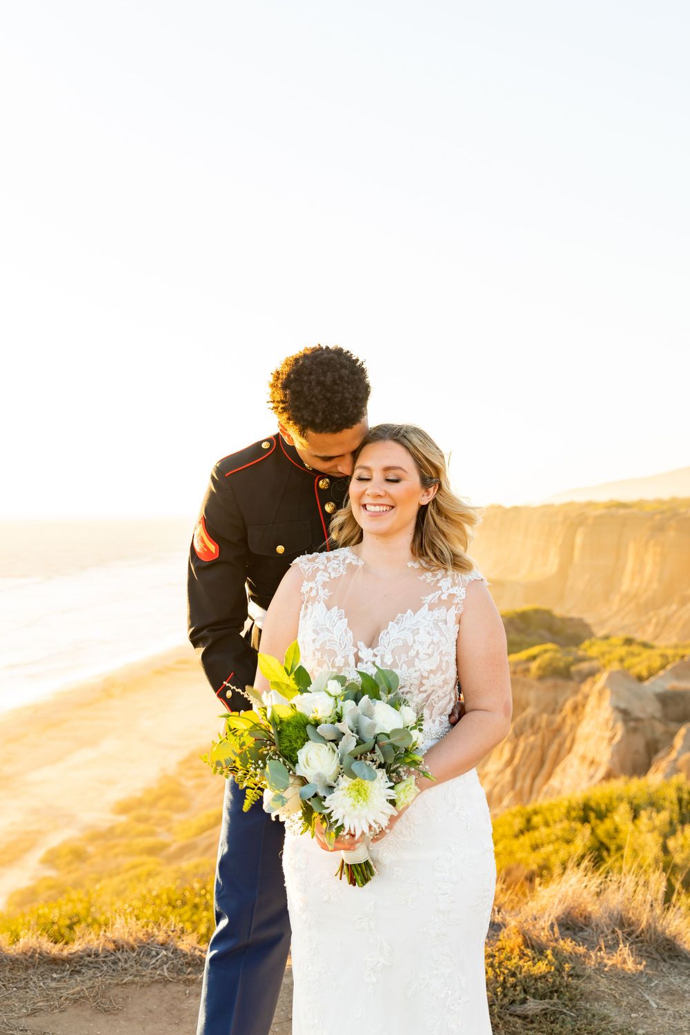 A couple in wedding attire embraces on a coastal cliff during golden hour with a beautiful bouquet of flowers.
