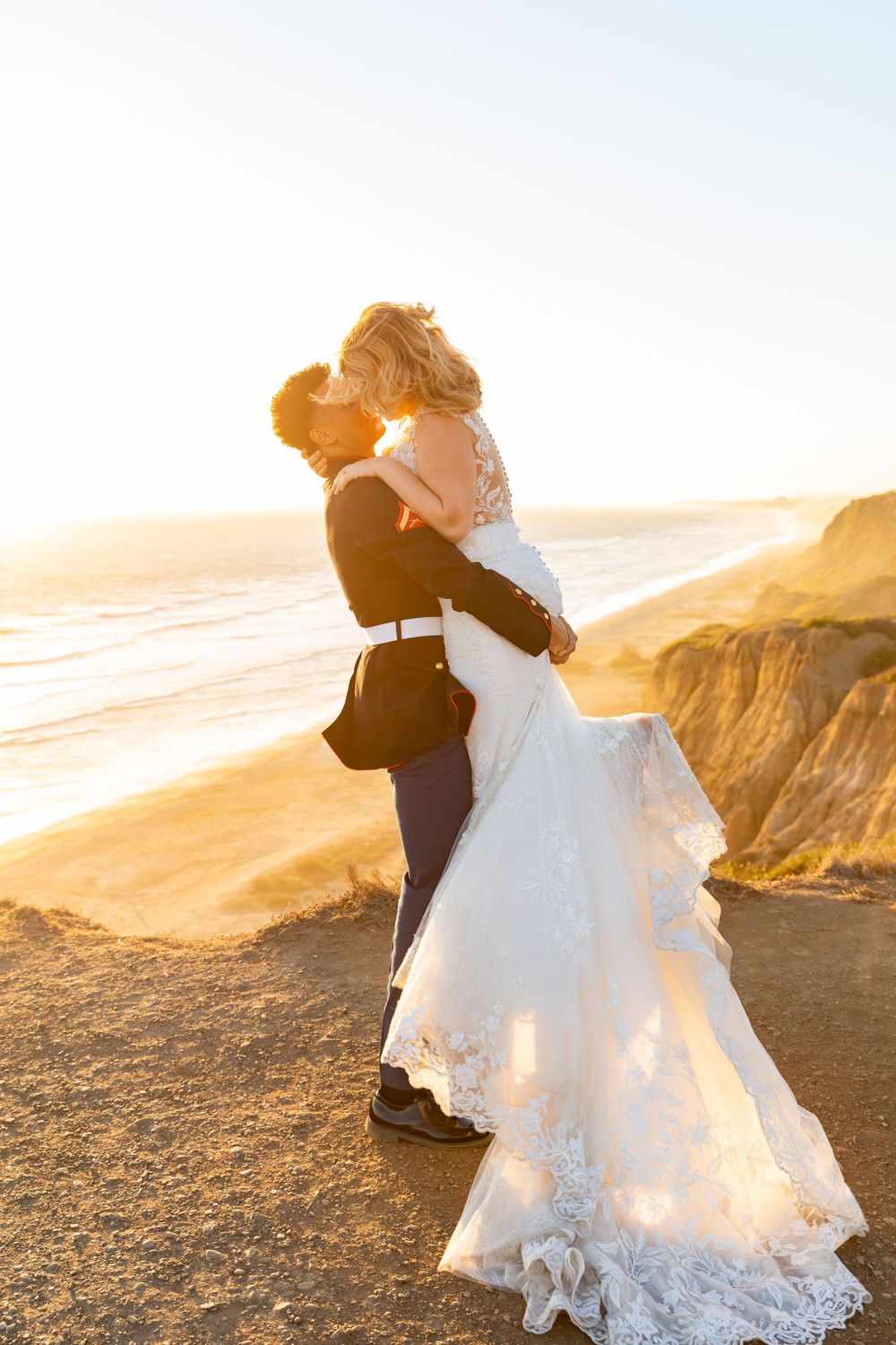 Newlyweds share an intimate moment on a clifftop with the sun setting over coastal cliffs.