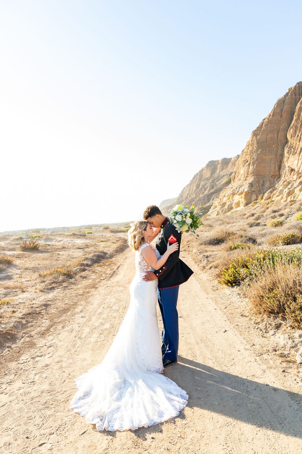 A military service member and bride embrace on a desert path with dramatic cliffs in the background.