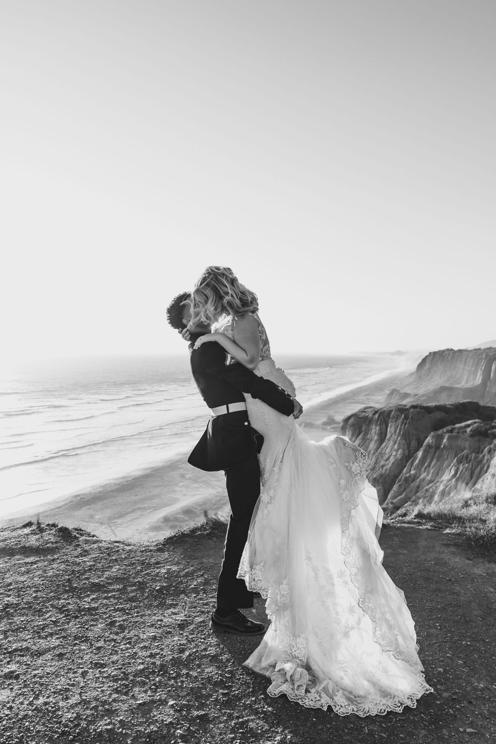 A black and white wedding photo showing an intimate embrace on a dramatic coastal cliff.