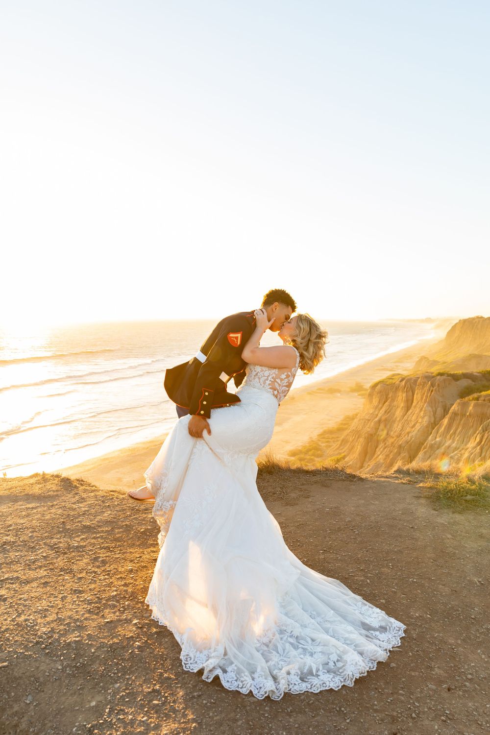 A tender dip kiss between newlyweds silhouetted against a golden sunset on coastal cliffs.