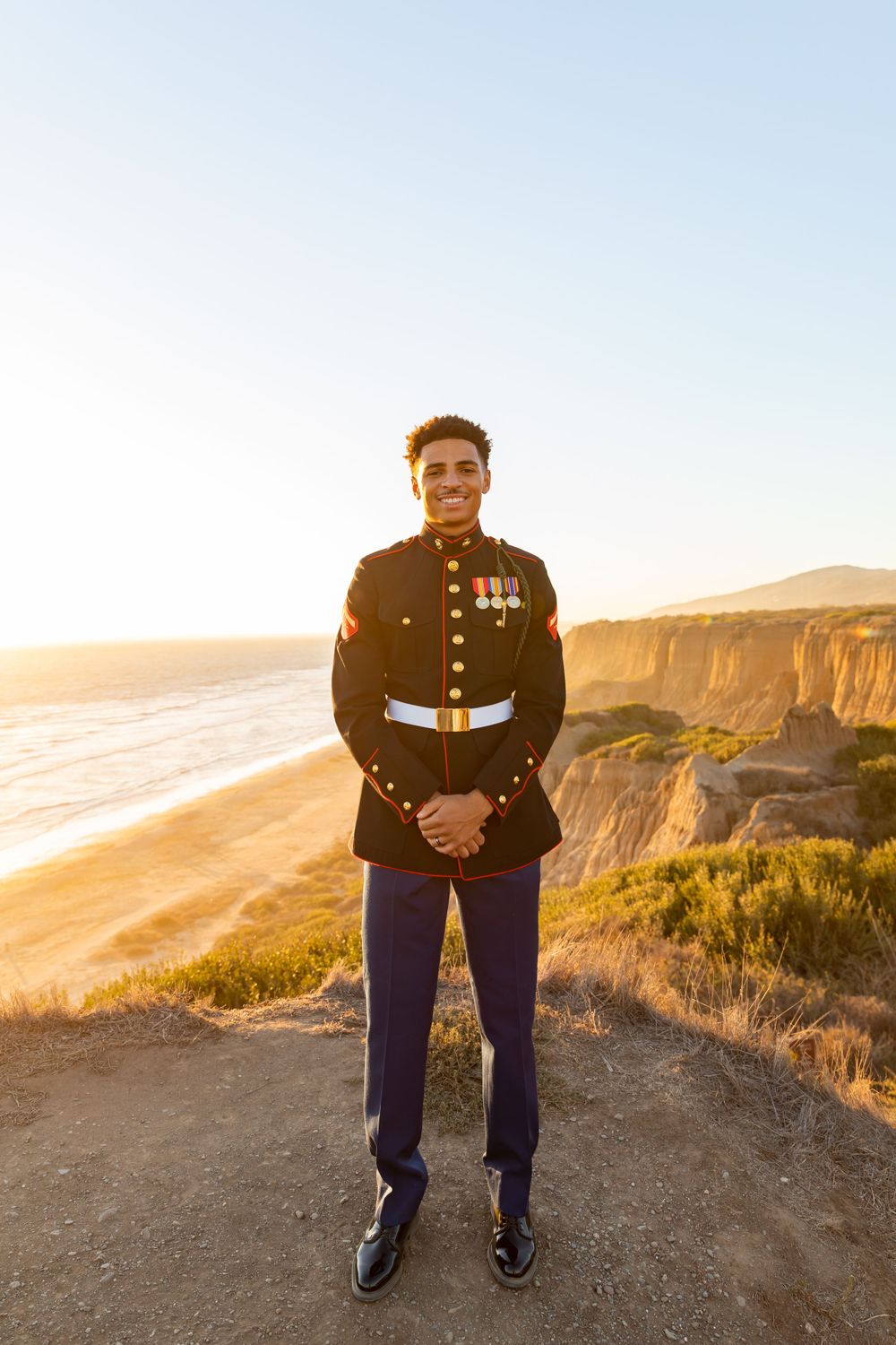 Marine in dress uniform standing at attention on coastal cliff at sunset.