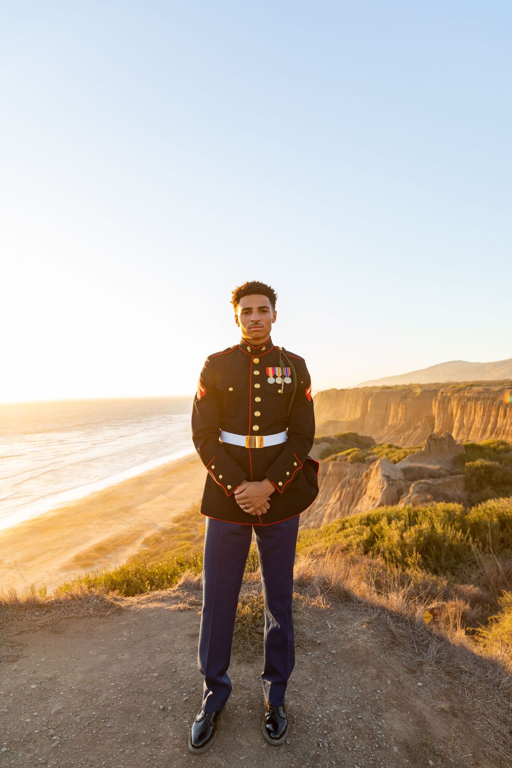 Marine in full dress uniform stands at attention on coastal cliff path.
