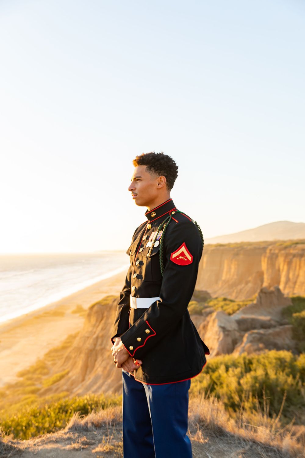 Marine in dress uniform gazes at sunset over ocean cliffs.