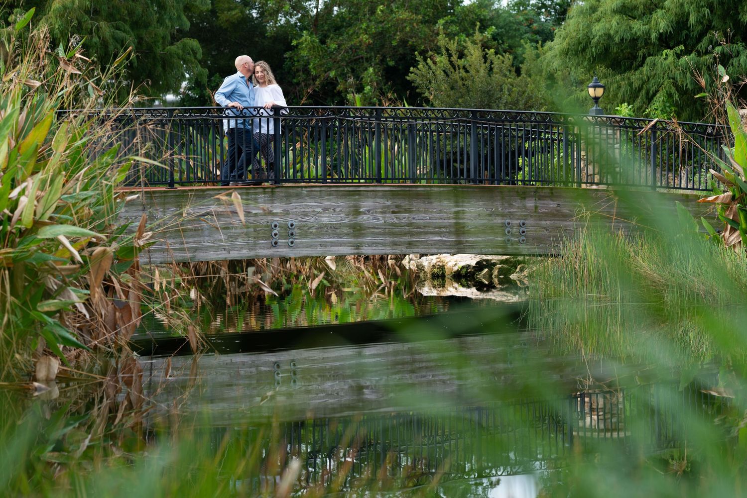 Couple stands on rustic bridge over pond surrounded by lush greenery.