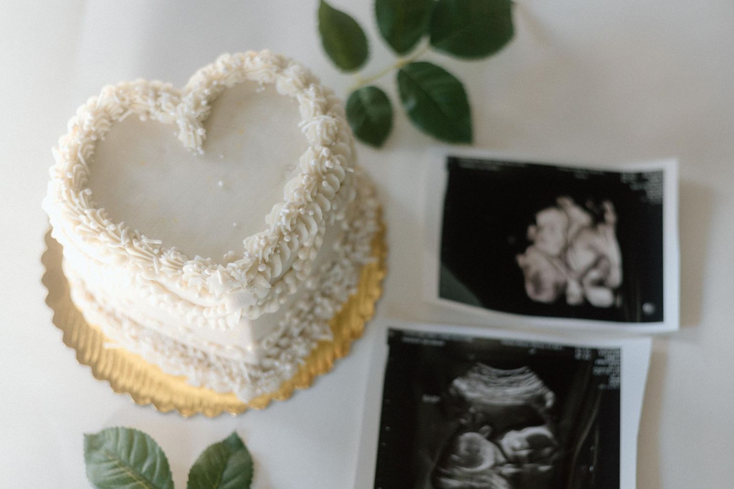 A heart-shaped white cake sits beside ultrasound photos and green leaves in a pregnancy announcement setup.