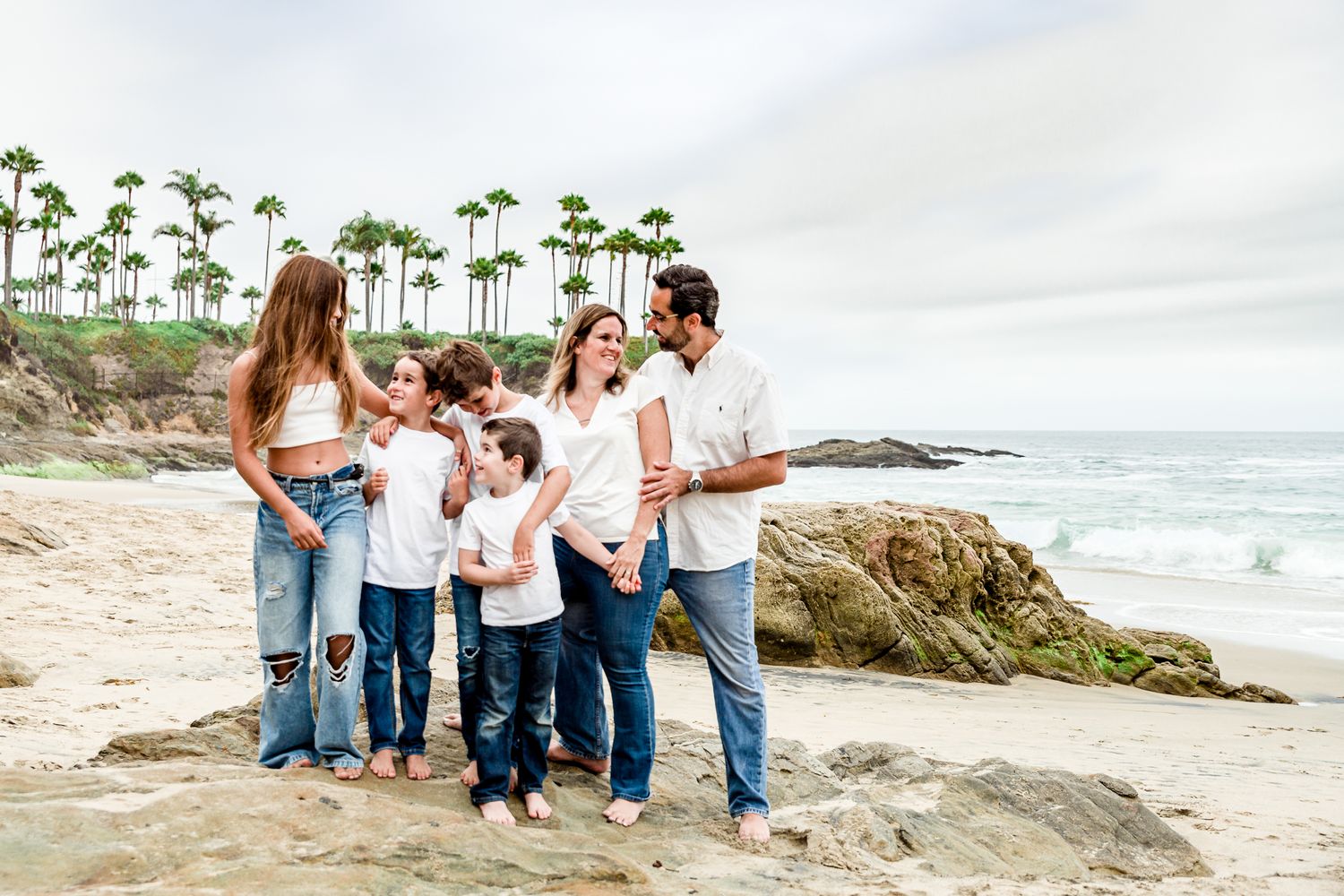 A family in coordinated casual outfits stands together on rocky coastal cliffs near the ocean by Stephanie Coulter Photography