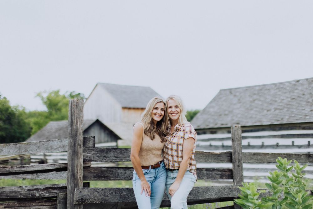 Two friends laughing together by wooden fence at rustic farm location.