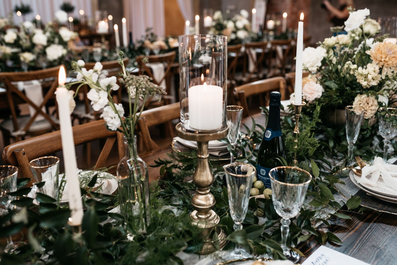 Elegant wedding table setting with white candles, crystal glassware and lush greenery garland on a wooden table.