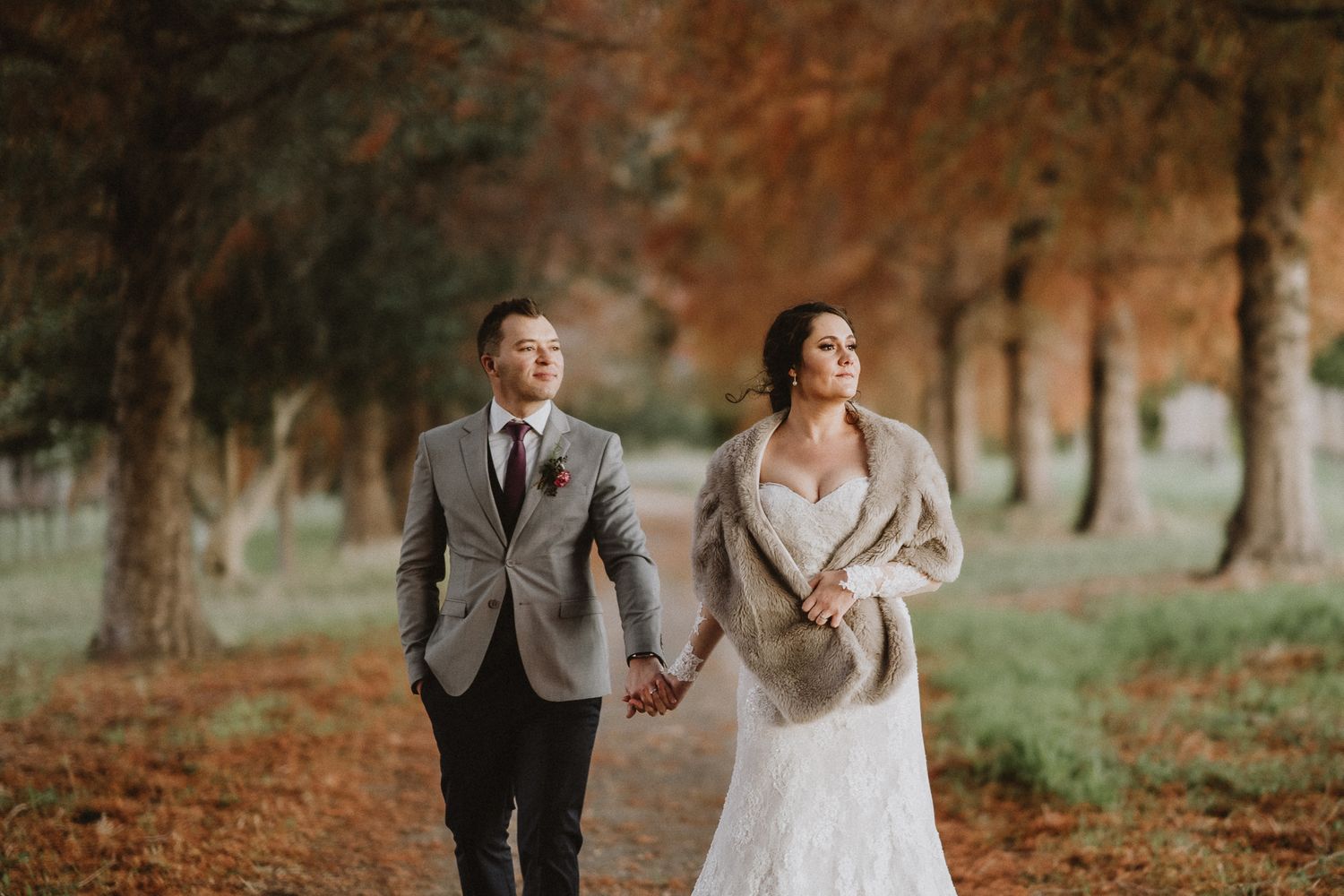 Newlyweds walk hand in hand through autumn trees with fallen leaves and warm lighting.