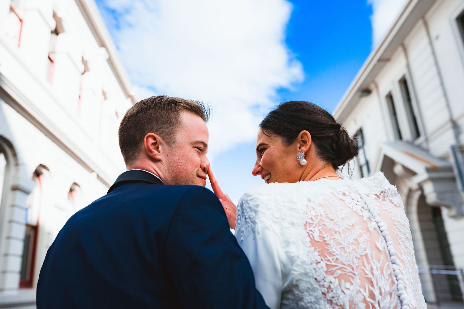 A couple in wedding attire shares an intimate moment against white architecture and blue sky.