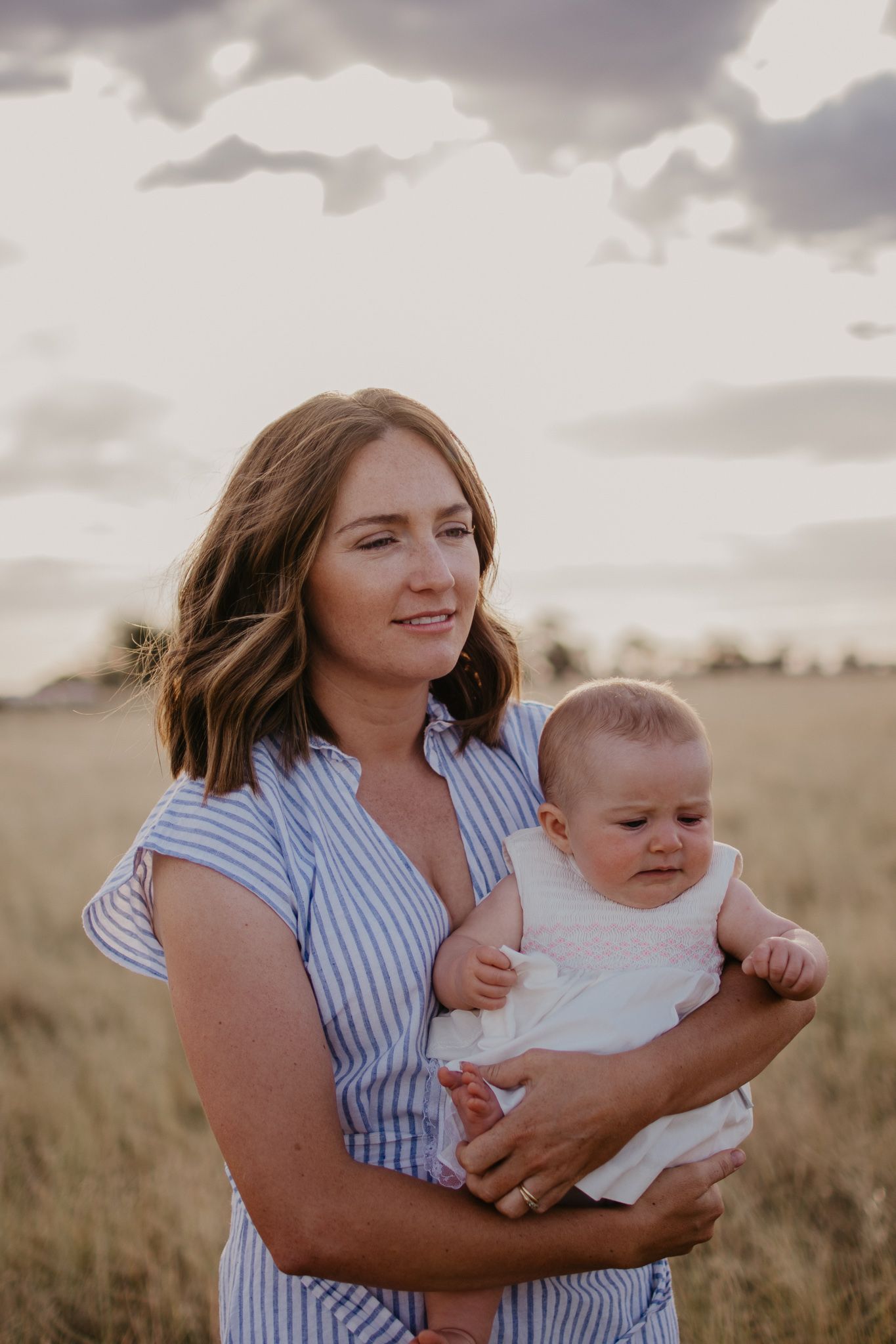 Strong country mum, in blue and white strip dress, standing in the paddock, looking out, holding baby