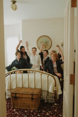 A celebratory group poses excitedly on a bed during wedding preparations.