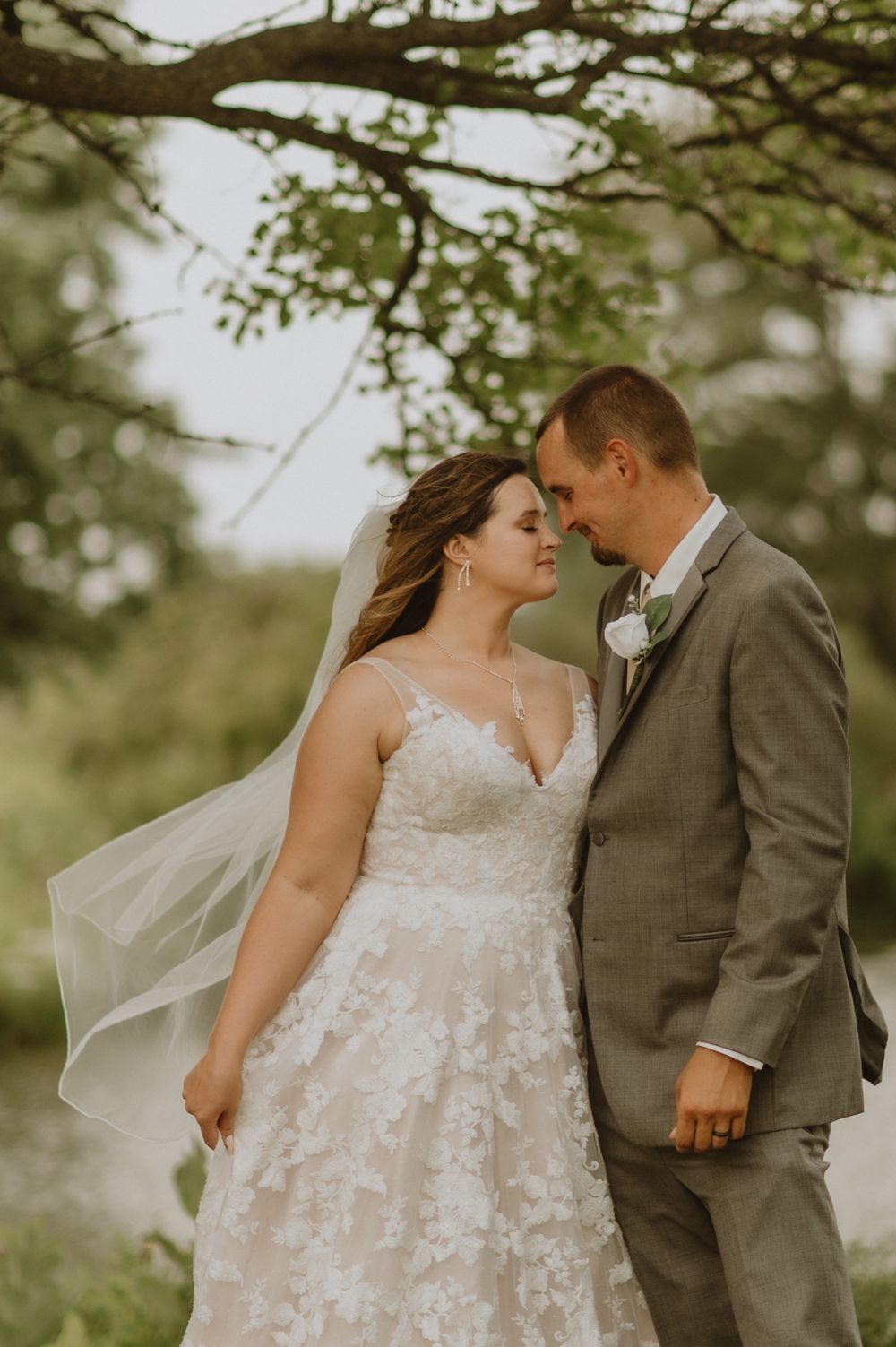 A romantic wedding portrait of a couple in formal attire beneath a leafy tree.