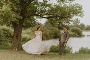 A bride twirls her lace dress while posing with her groom by a lakeside tree.