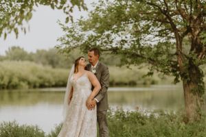 A romantic wedding portrait of a couple embracing by a serene lake under tree branches.