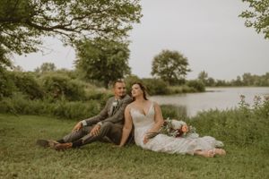 Newlyweds relaxing on grass by a serene lake with a bouquet of flowers beside them.