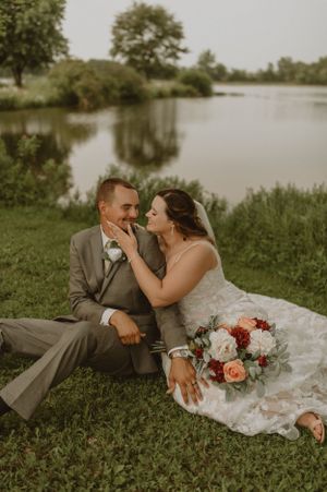 A tender moment between newlyweds sitting lakeside with a colorful floral bouquet.