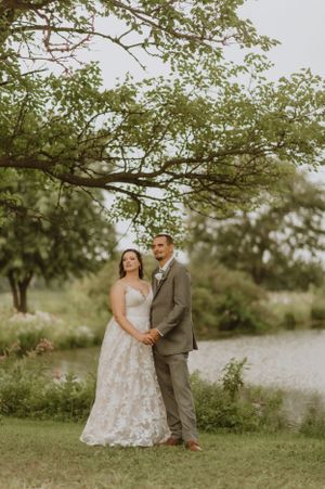 A wedding couple poses together on grass beneath a sprawling tree by a lake.