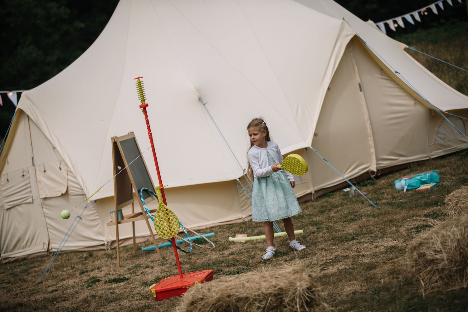 A child in a blue dress plays outside large canvas bell tents at an outdoor event.