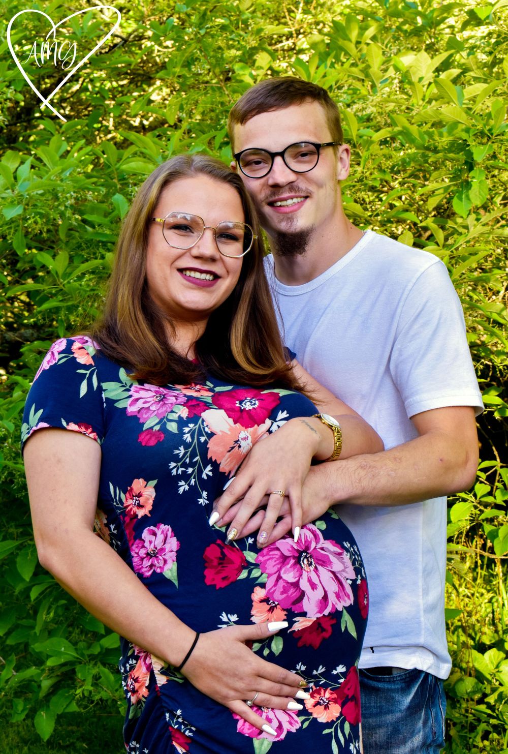 Pregnant couple outdoors, woman holding baby bump, man in white t-shirt embracing her, green foliage background. Maternity Photographer.