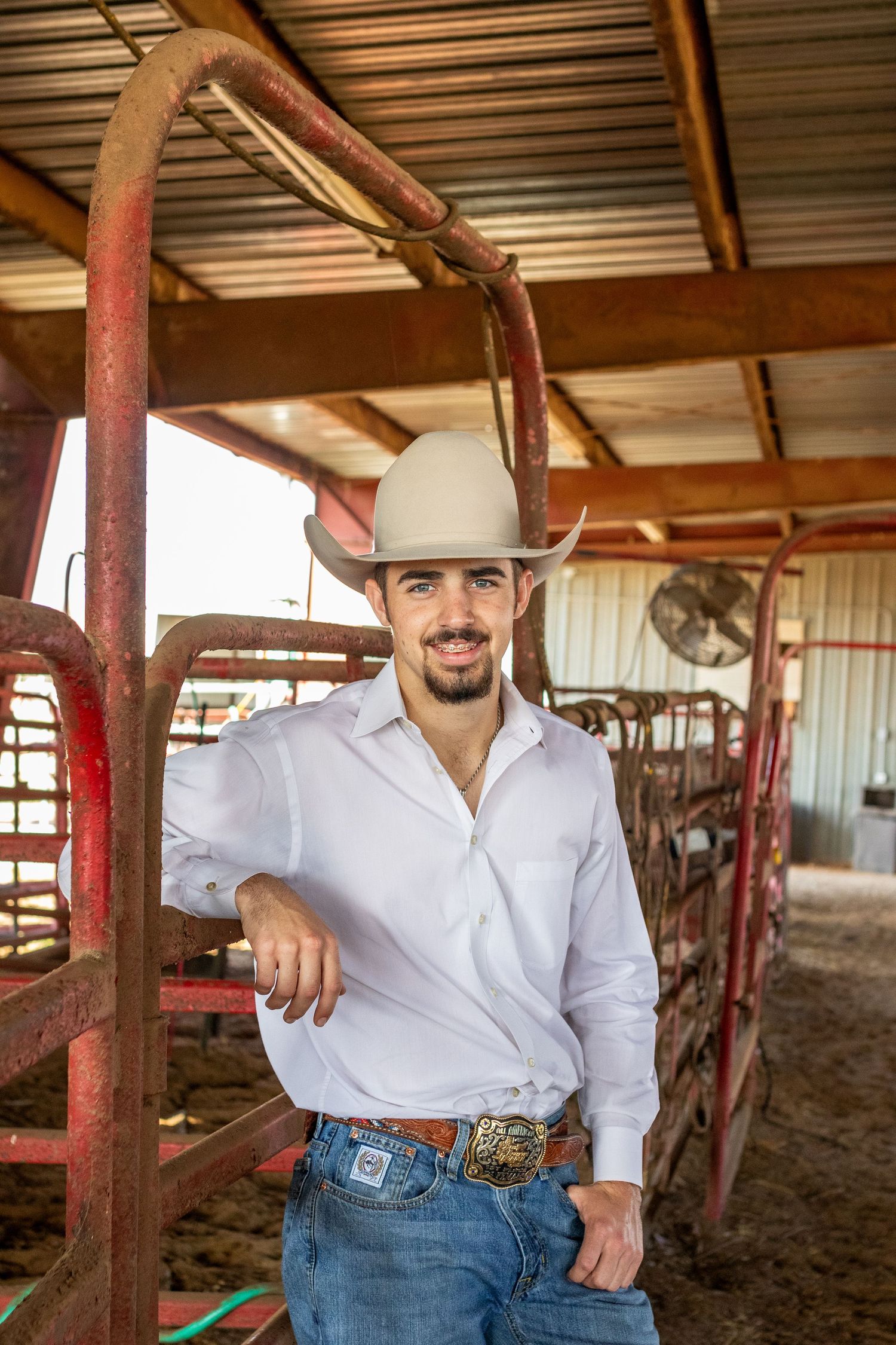 Handsome young man in white shirt and cowboy hat leans on red cattle pen in barn.