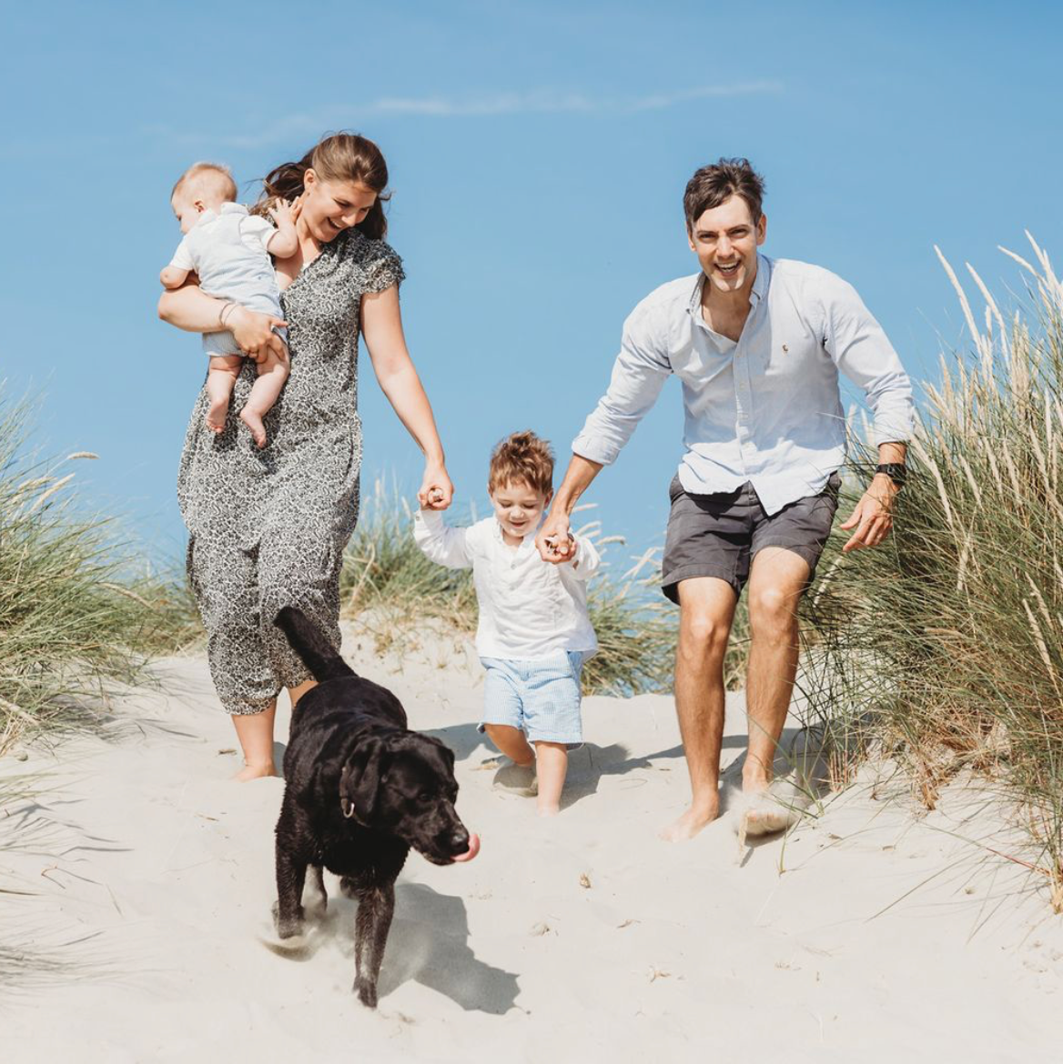 Family and black dog running together up sandy beach dunes surrounded by tall grass during a family beach photoshoot