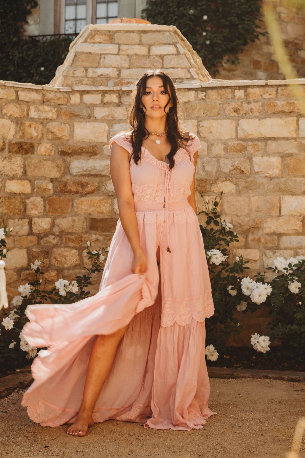 Romantic pink flowy dress against stone wall backdrop with white roses in garden setting.