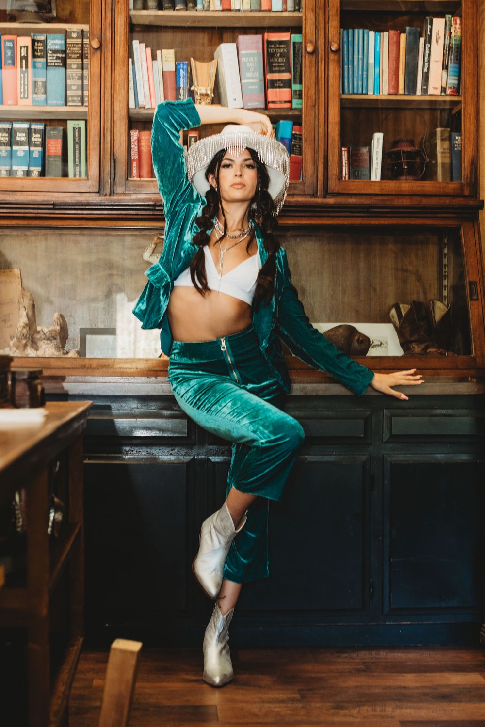 Dynamic poses in emerald velvet outfit against vintage library bookshelf backdrop.