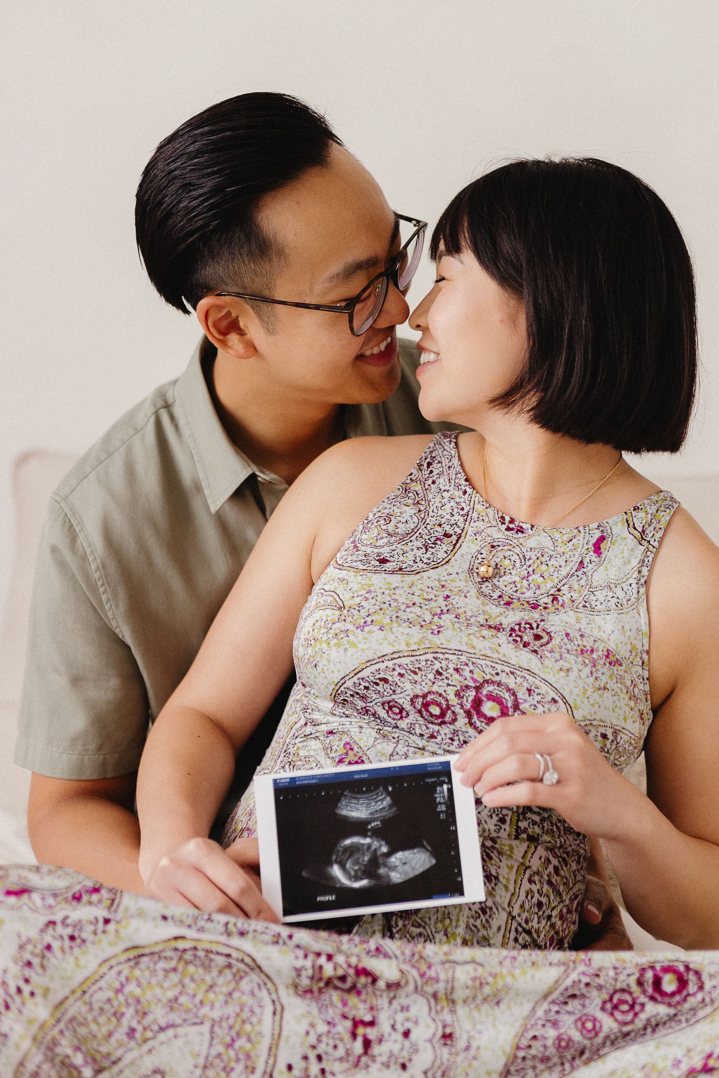 An expecting couple poses with ultrasound photos while wearing casual clothing against a white background.