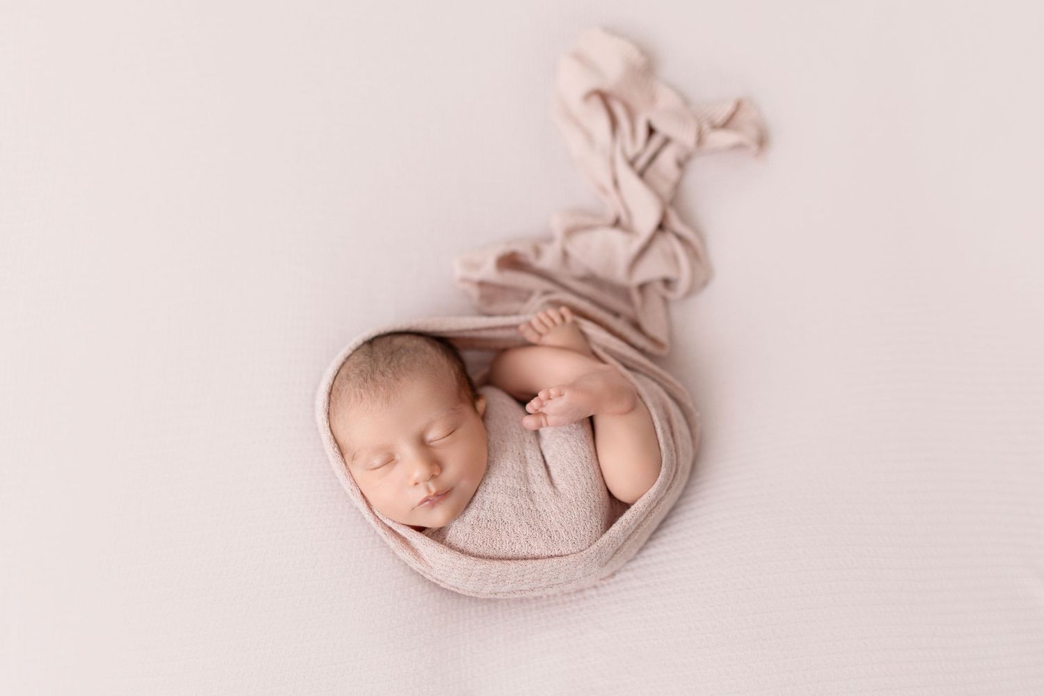 Newborn baby sleeping peacefully in a round bowl with soft pink fabric and bow decoration.
