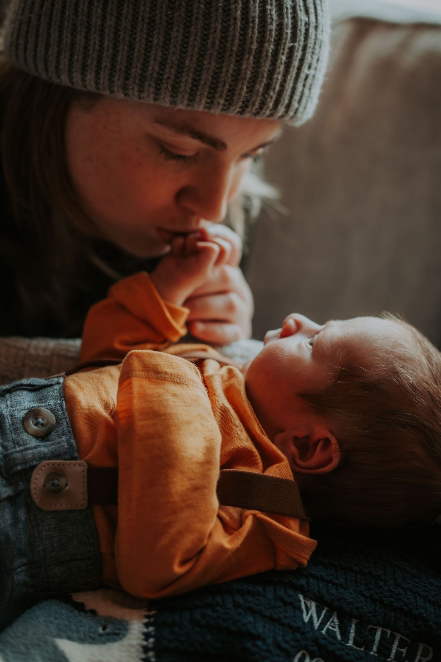 A woman in a gray knit beanie kisses the tiny hand of a swaddled newborn baby lying on a cozy couch.