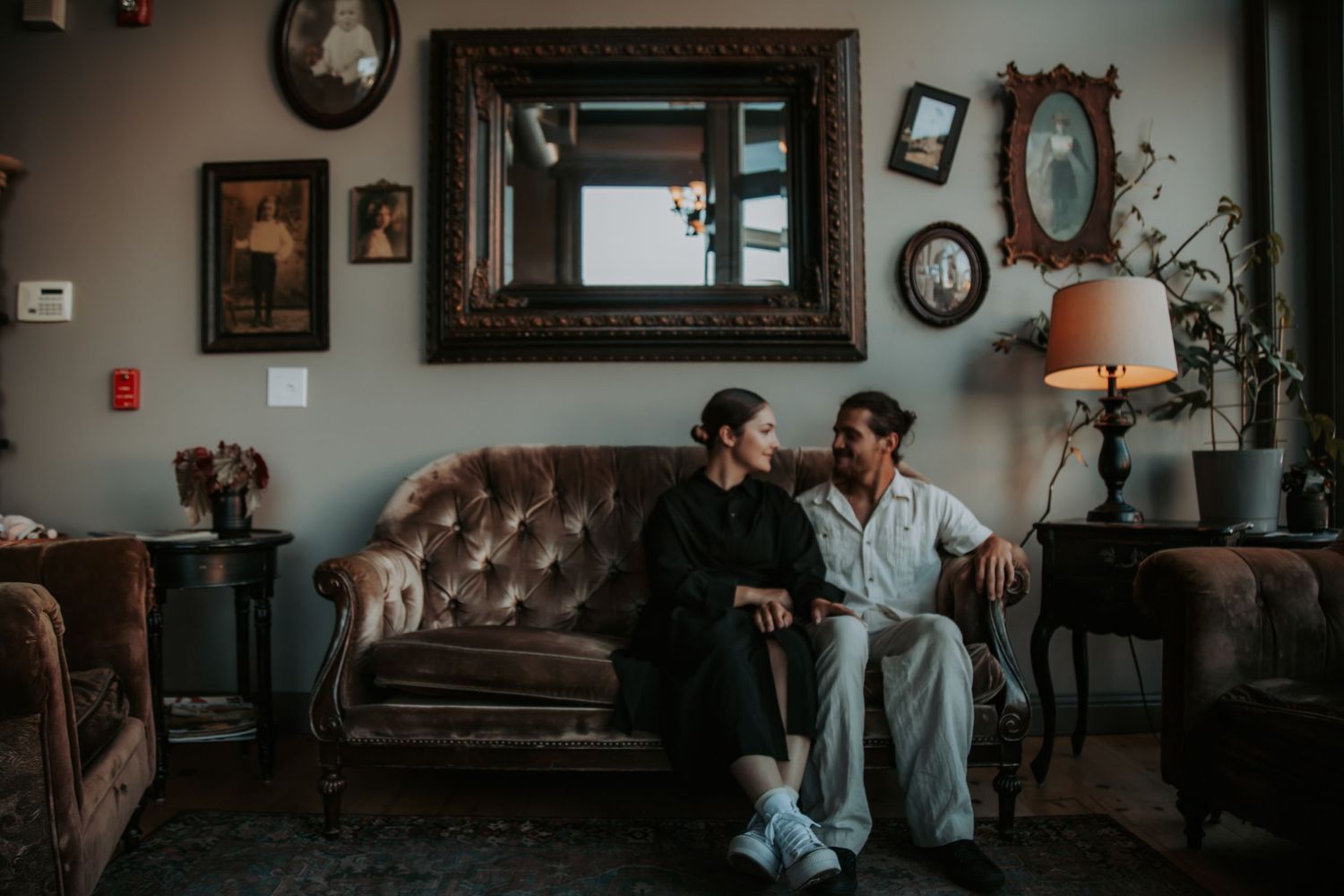 Couple in matching dark and light loungewear sit close together on a tufted leather sofa in a vintage-styled living room.