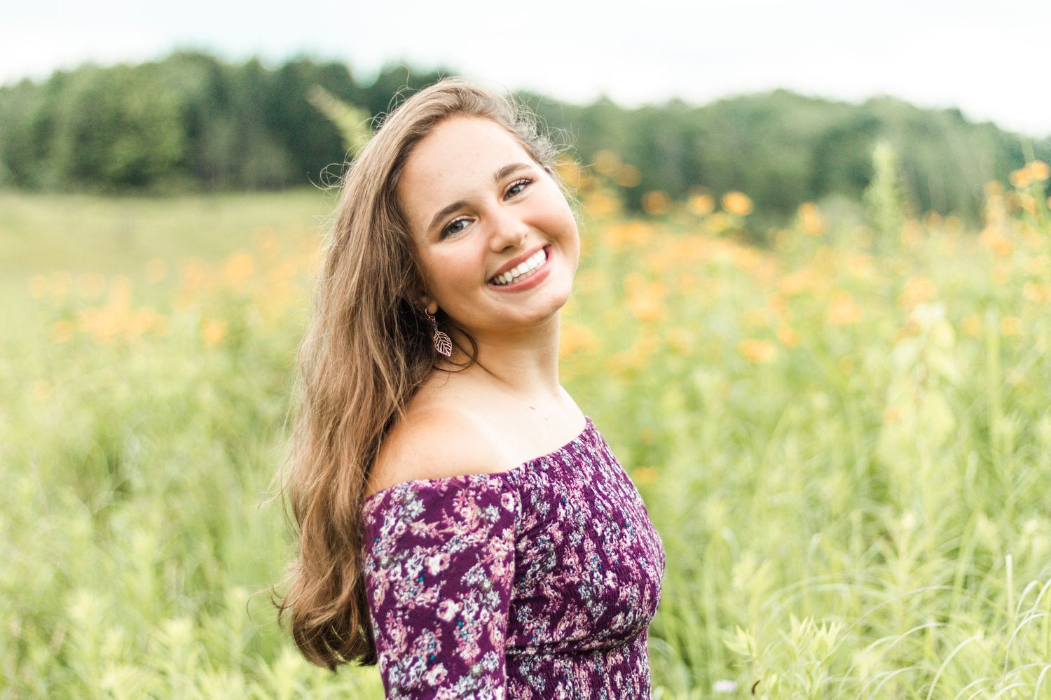 A person in a purple floral dress poses in a sunny meadow with yellow wildflowers.