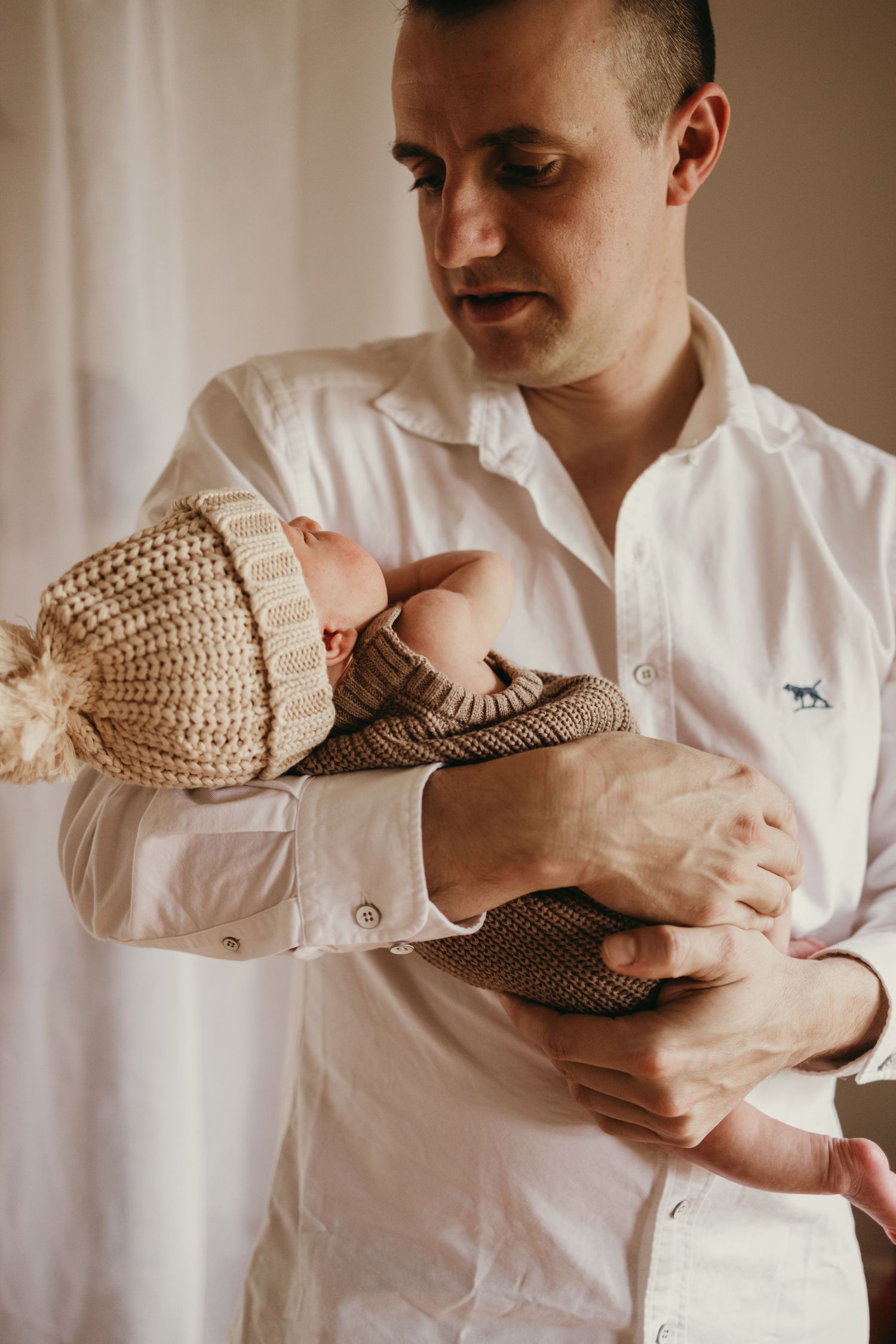 Parent holding sleeping newborn wearing knitted beanie and sweater in warm light.